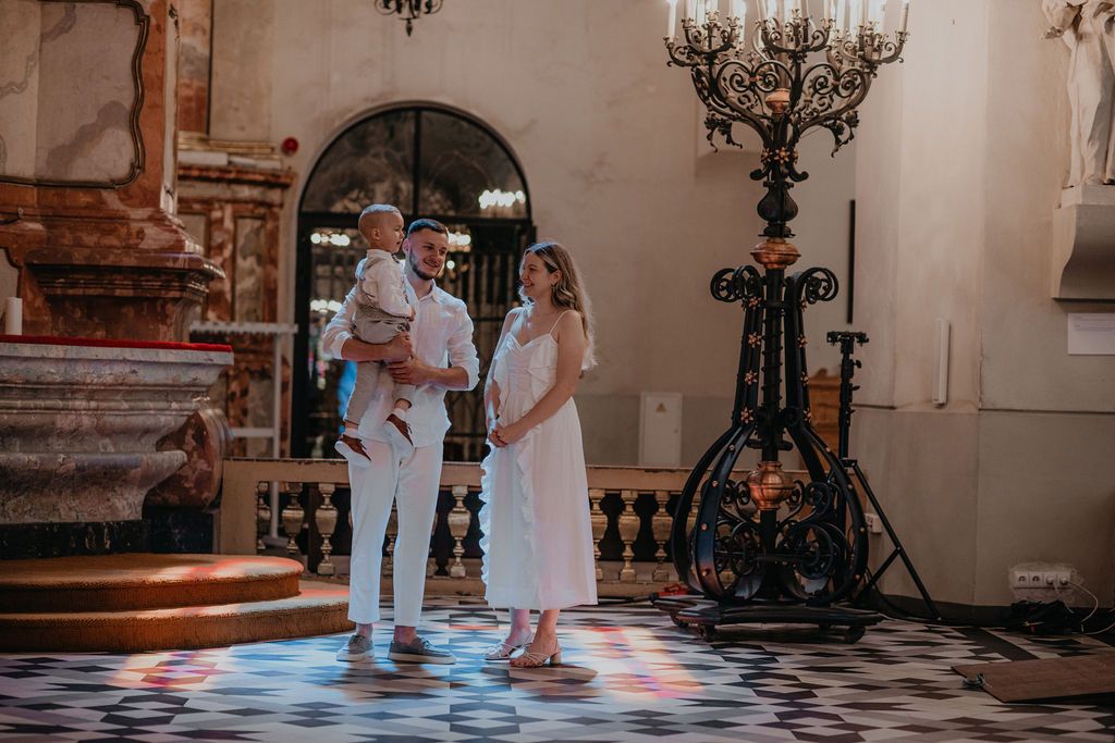 Family in white attire during a baptism ceremony inside an ornate church with decorative chandeliers.