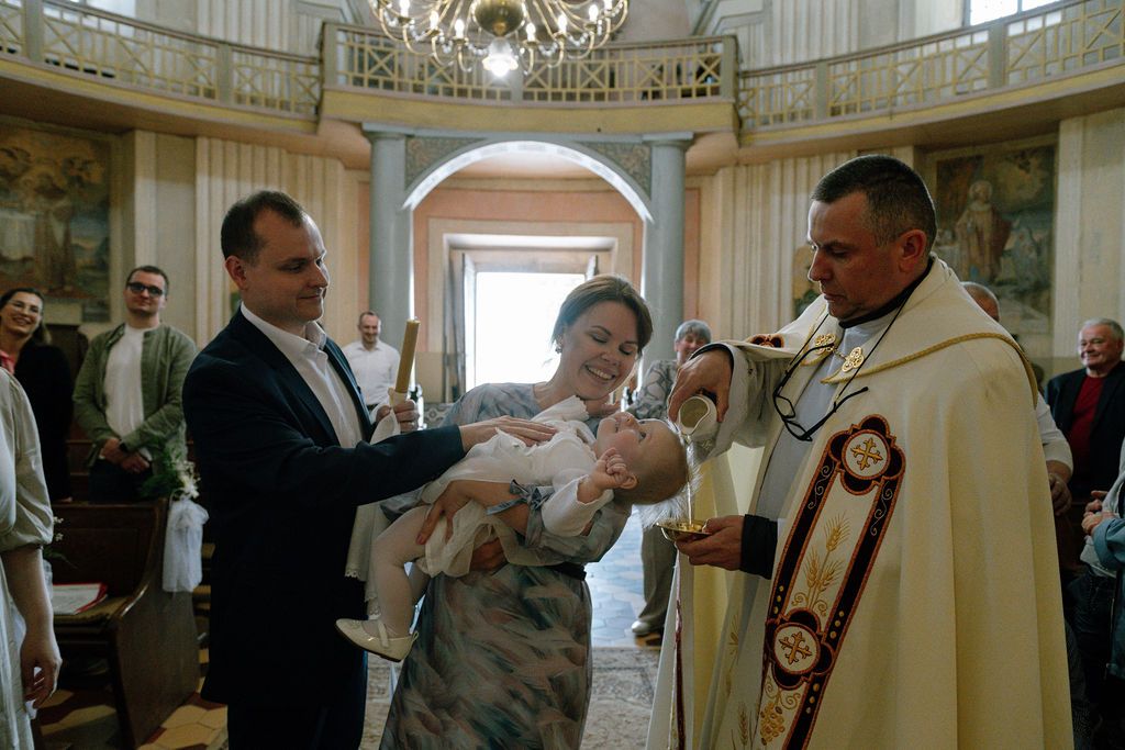 Catholic priest performing a baptism ceremony on an infant held by parents in an ornate church.