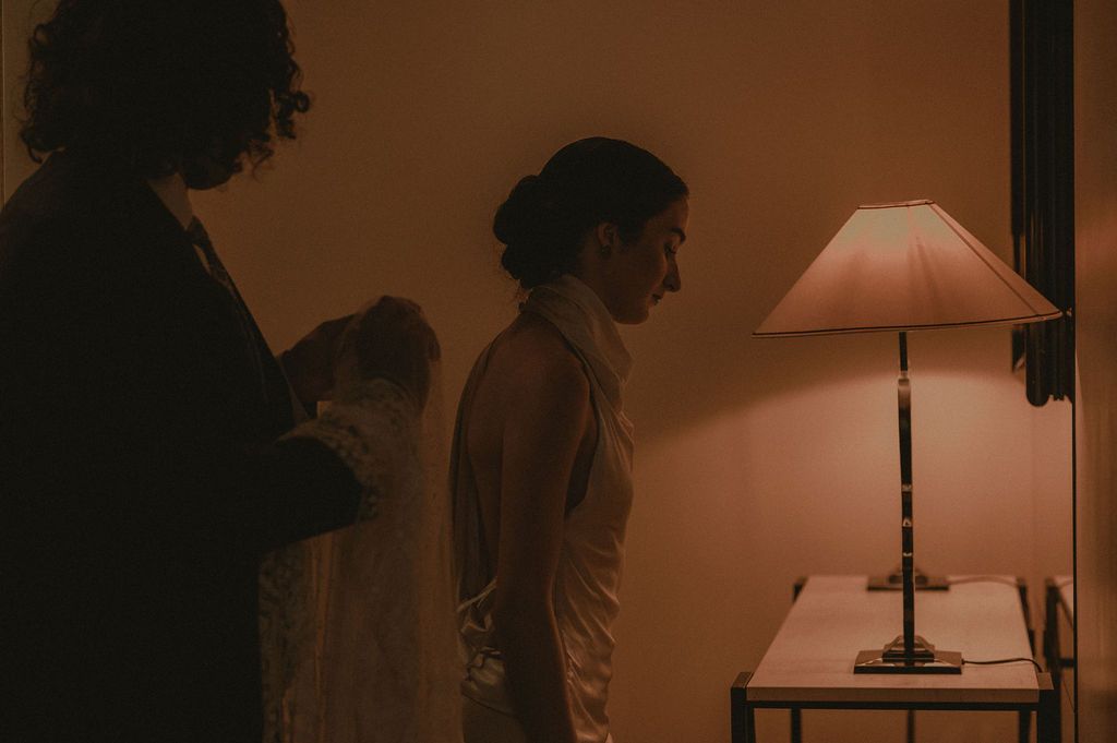 Two women in elegant attire stand in a warmly lit room beside a glowing table lamp in a moody, intimate setting.