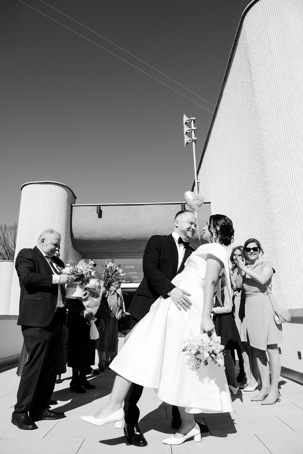 Black and white photo of a bride and groom sharing a kiss outside a modern building surrounded by wedding guests.