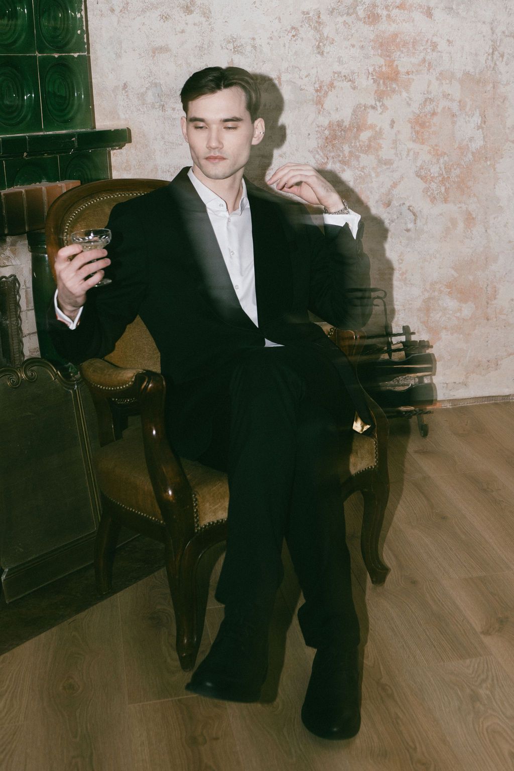 Sophisticated man in black tuxedo sitting in ornate chair holding whiskey glass in classic interior.