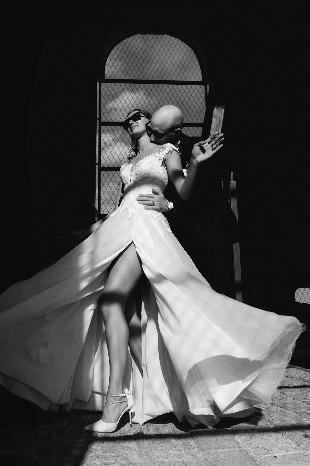 Black and white photo of a bride in flowing wedding dress and sunglasses posing dramatically with a bald man outdoors.
