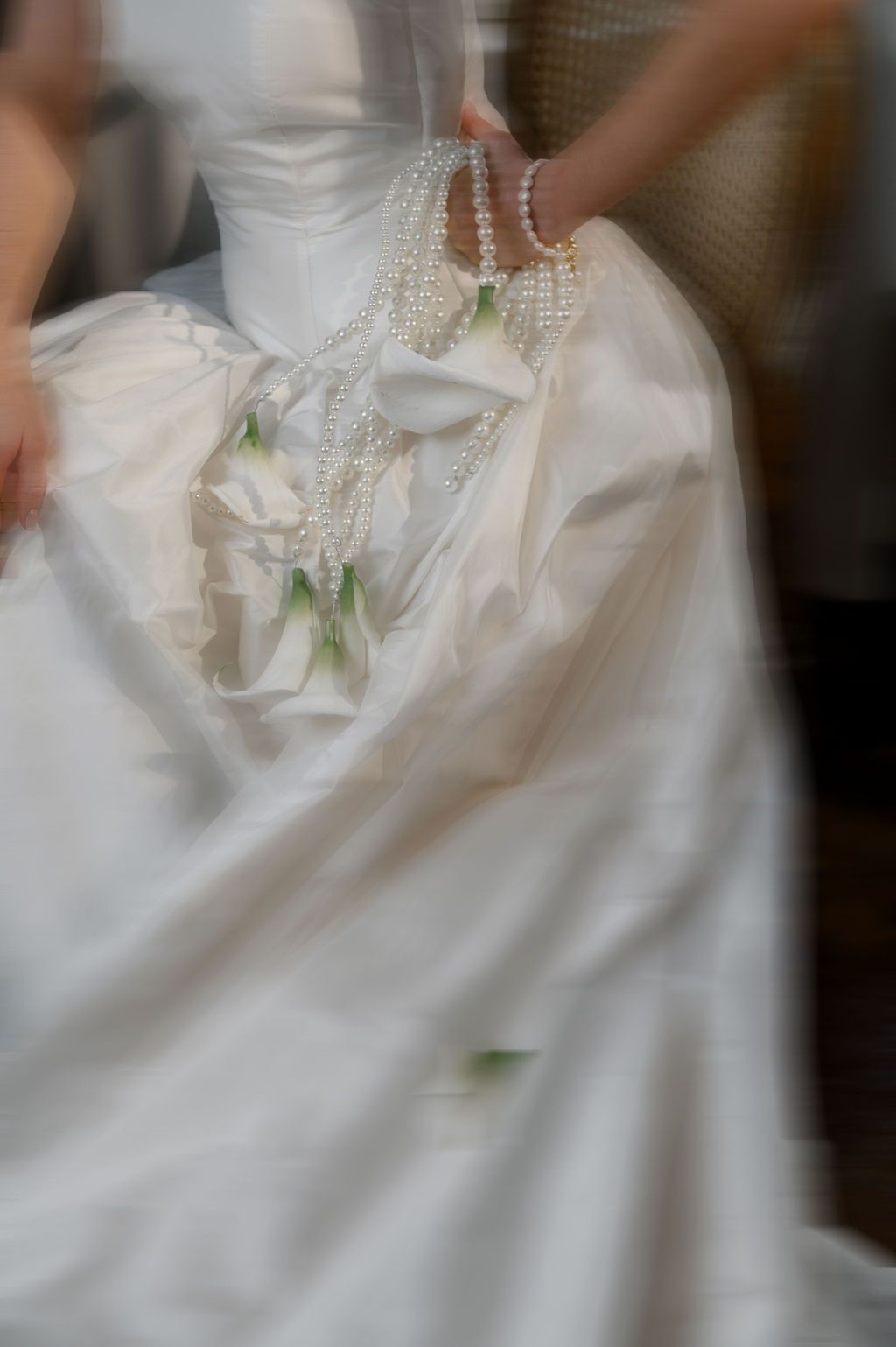 Close-up of bride's white satin wedding gown adorned with cascading pearl strands and delicate white floral bouquet.