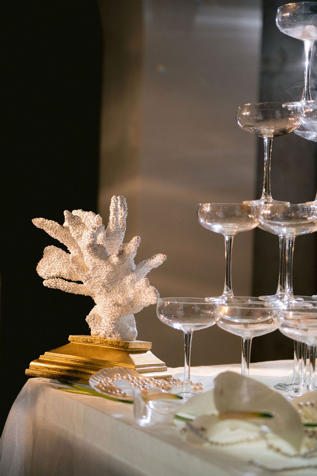 Elegant champagne tower with crystal coupe glasses beside a white coral sculpture on gold stand at a wedding reception.