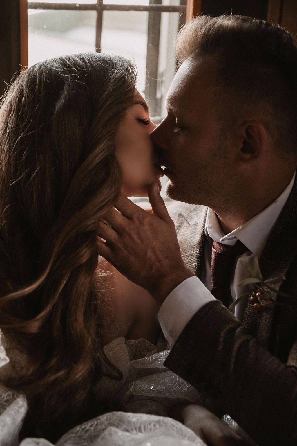 A bride and groom share a romantic kiss near a window in warm, moody lighting on their wedding day.
