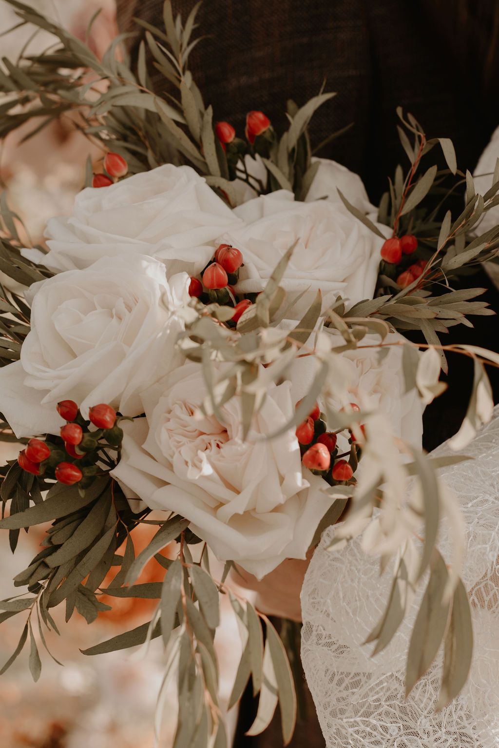 Close-up of a bridal bouquet featuring white roses, red berries, and olive branches held by a bride in lace.
