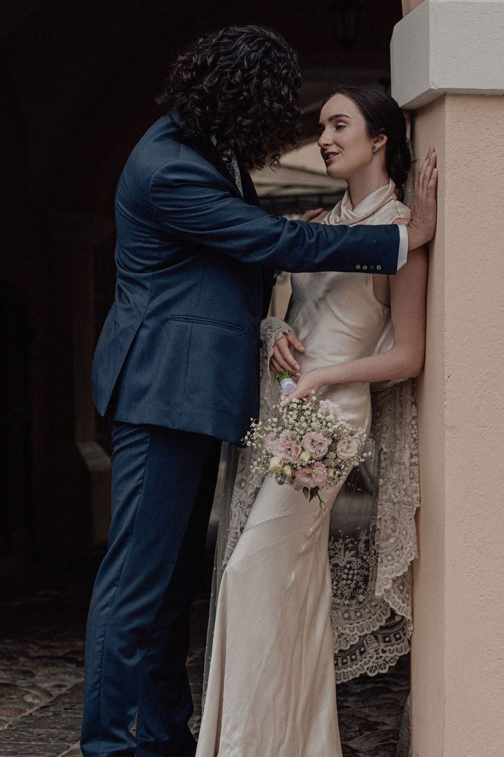 Bride in silk gown with lace veil holds pink bouquet as groom in navy suit leans in romantically against wall.