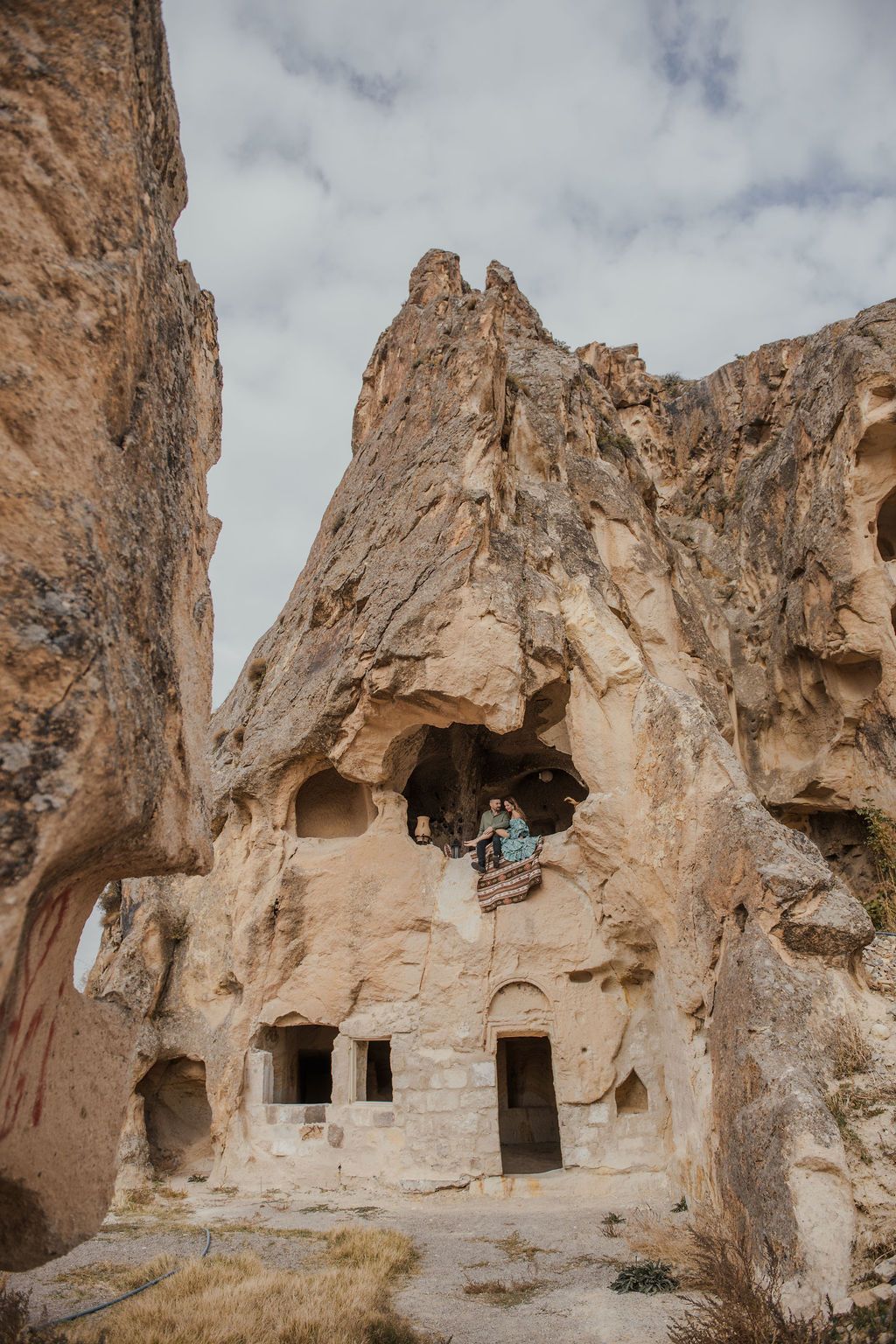People exploring ancient cave dwellings carved into towering rock formations in Cappadocia, Turkey, under a cloudy sky.