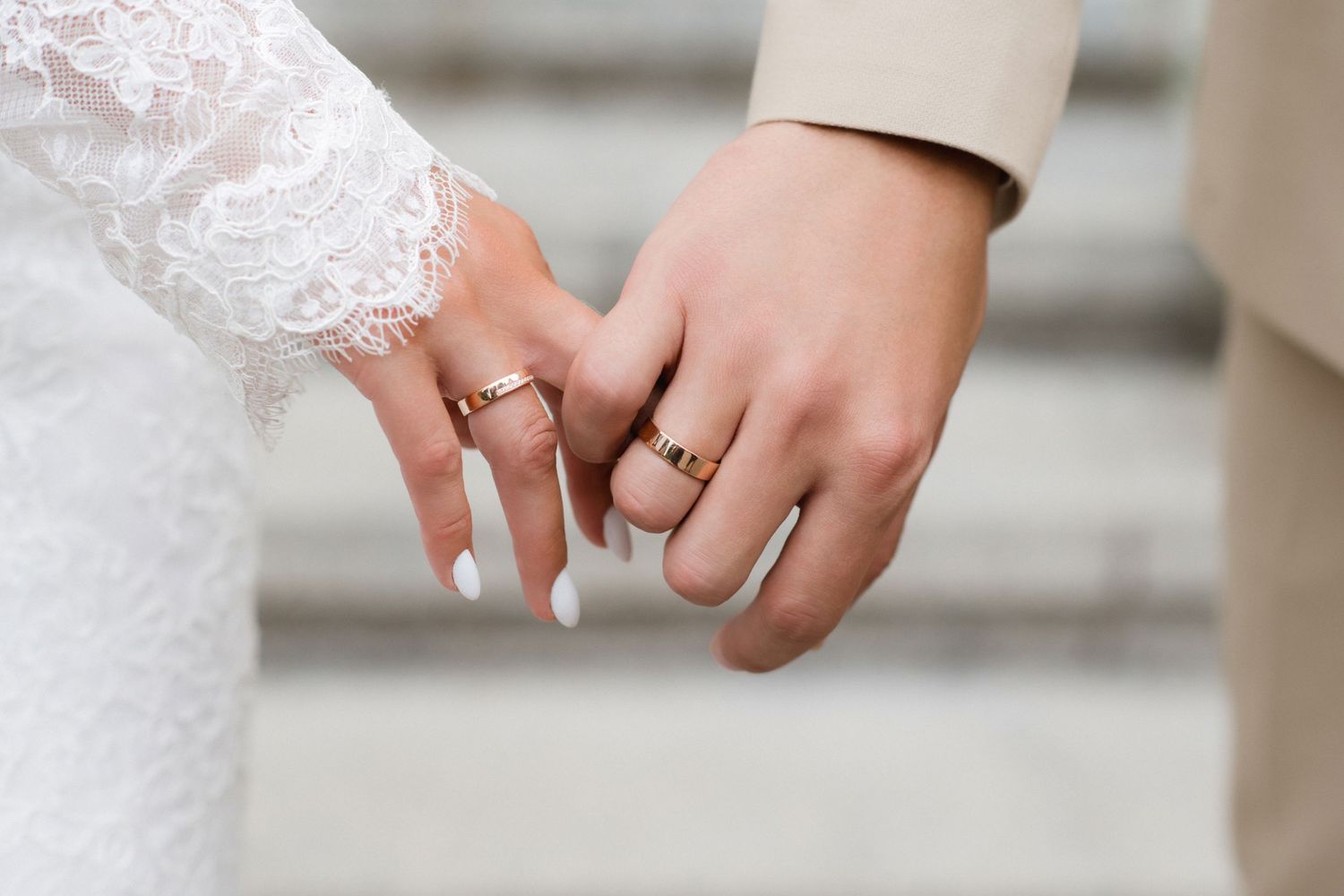Bride and groom holding hands showing gold wedding rings, bride wearing lace sleeve with white manicure.