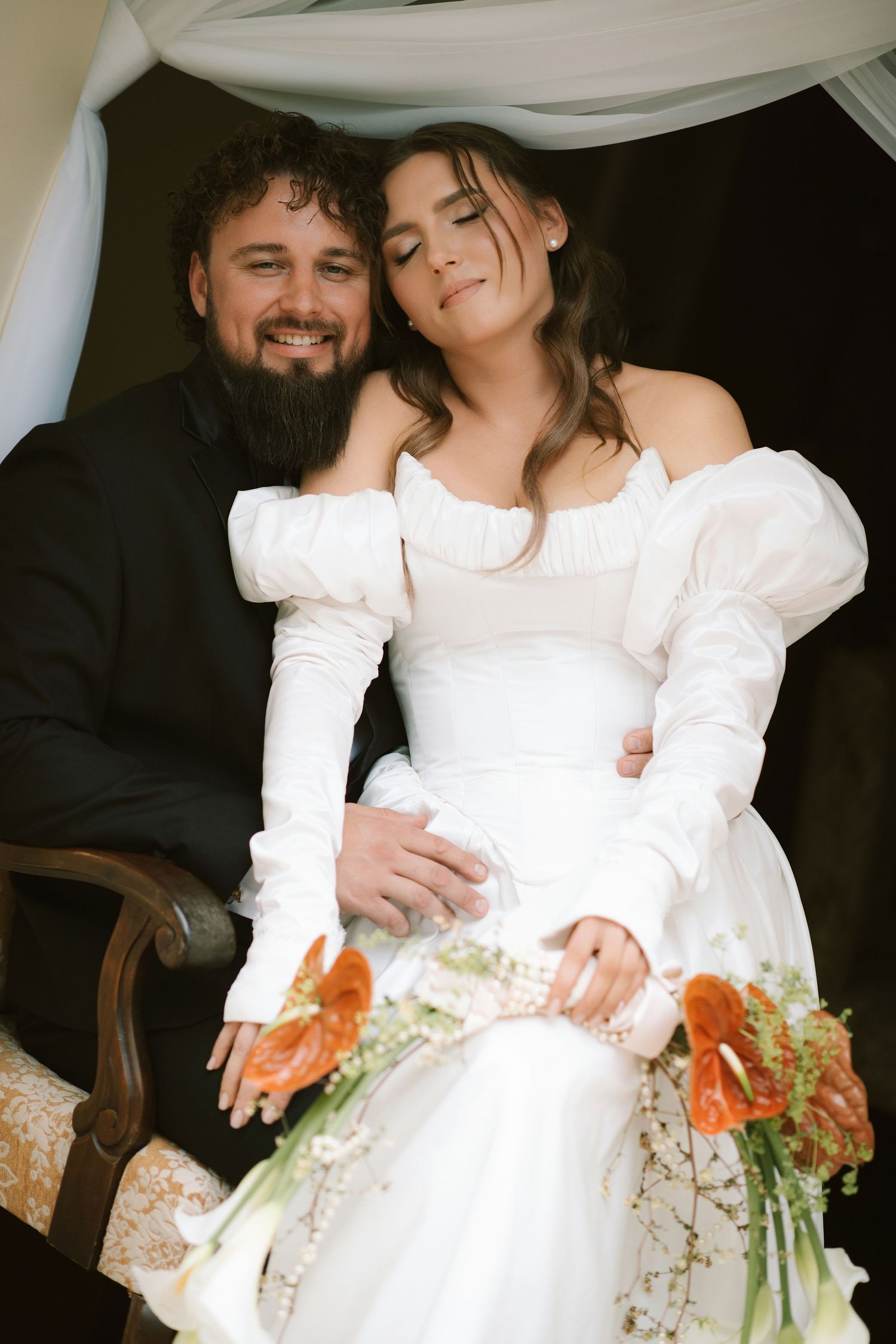 Bride in white off-shoulder gown holds orange bouquet while groom in black suit embraces her at wedding ceremony.
