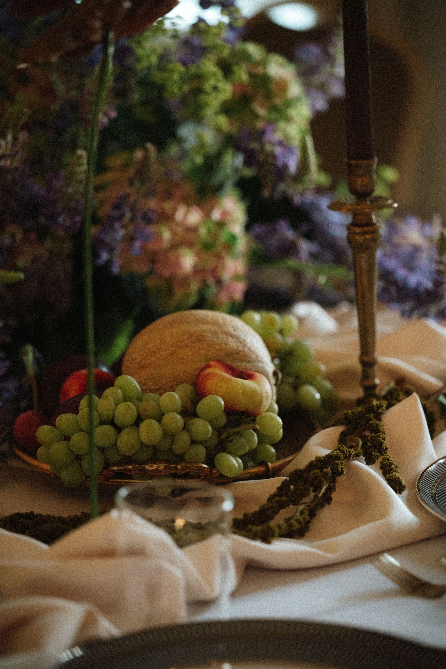 Elegant wedding table centerpiece with green grapes, cantaloupe, and peaches on a silver stand beside a brass candlestick.