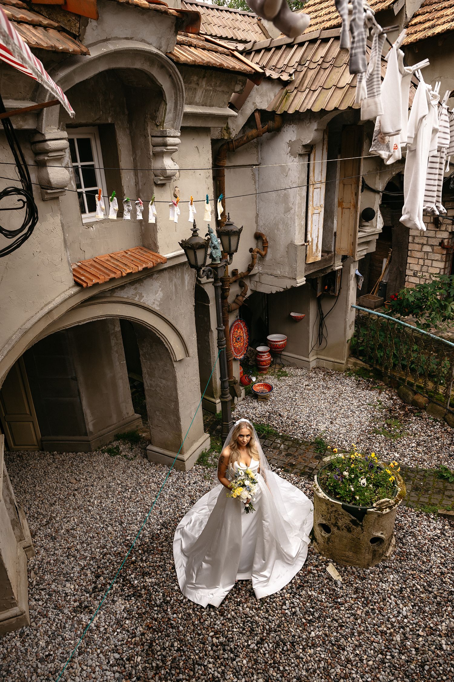 A bride in a flowing white dress stands in a rustic courtyard with stone walls and terracotta roofing.