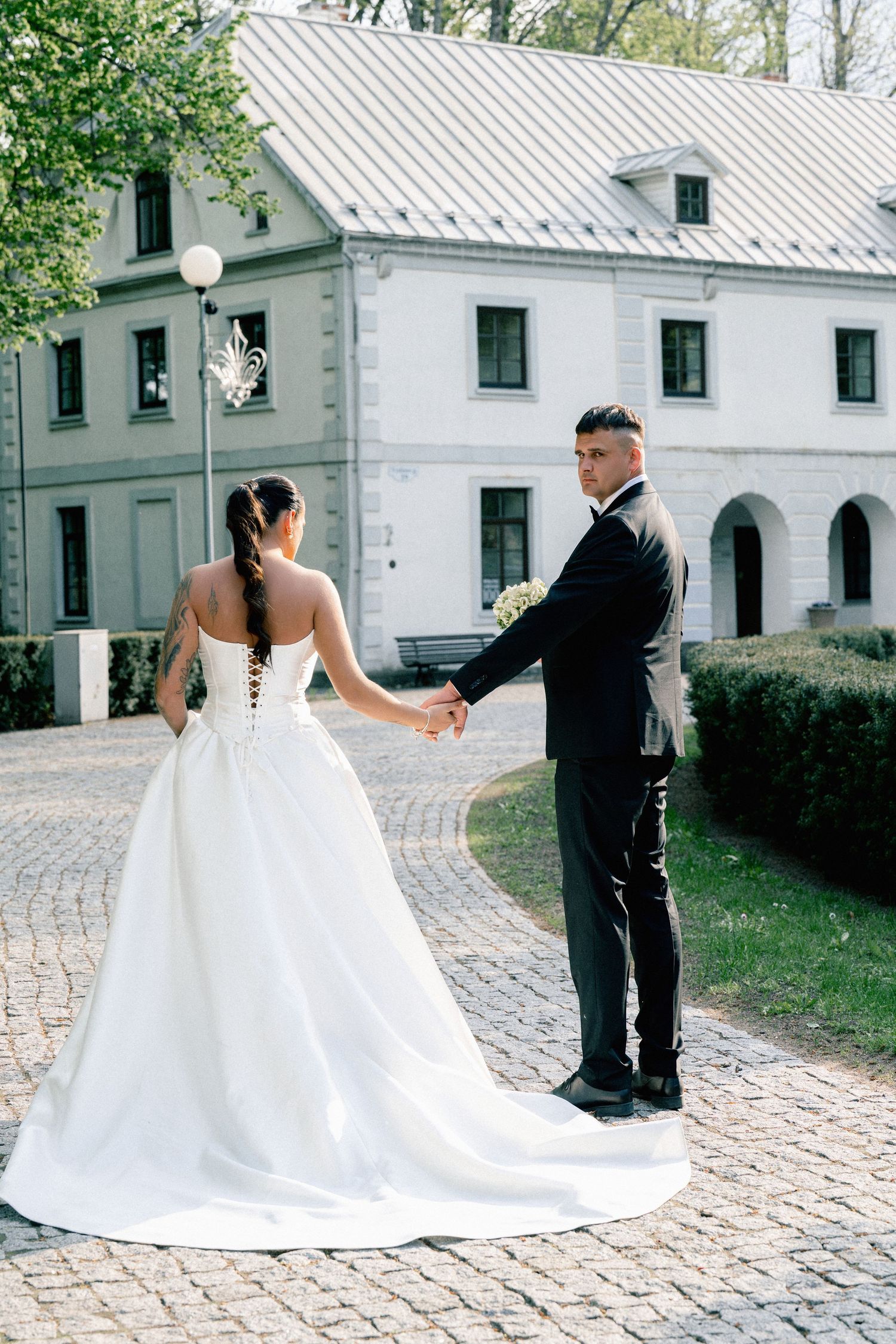 A bride in a white gown and groom in a black suit hold hands on a cobblestone path outside an elegant white estate.