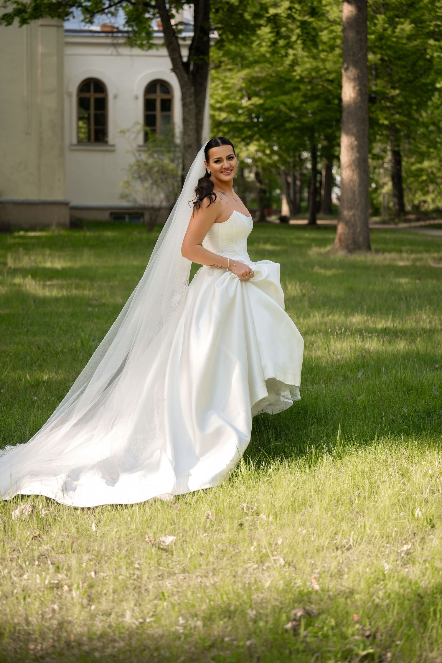 Smiling bride in a flowing white strapless gown and long veil poses gracefully in a lush green park setting.