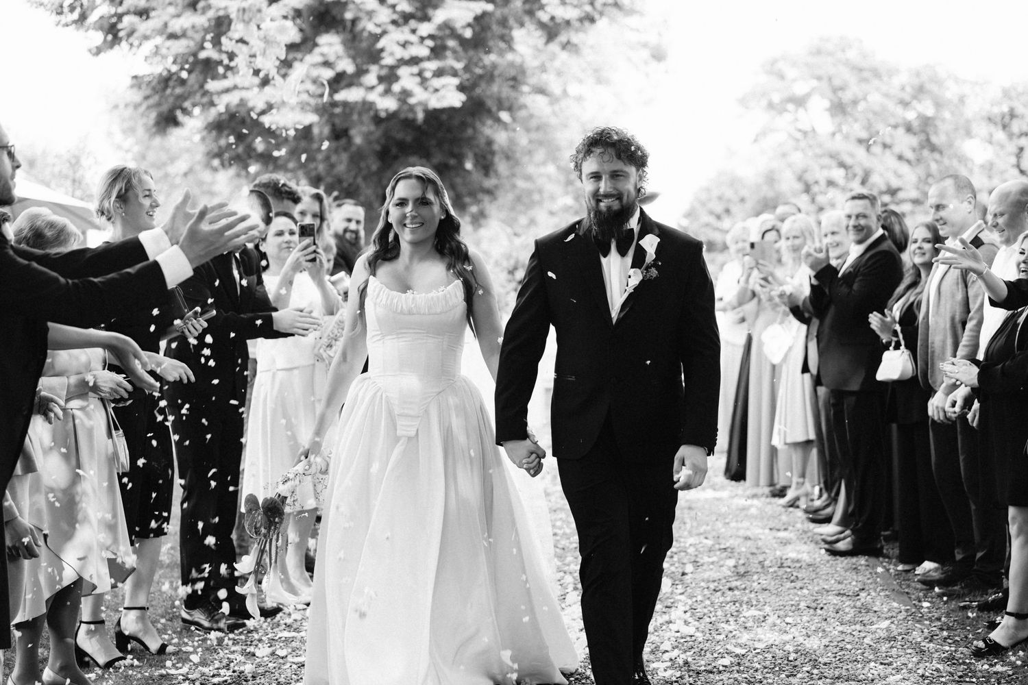 Black and white photo of newlyweds walking through confetti shower with smiling wedding guests lining the path.