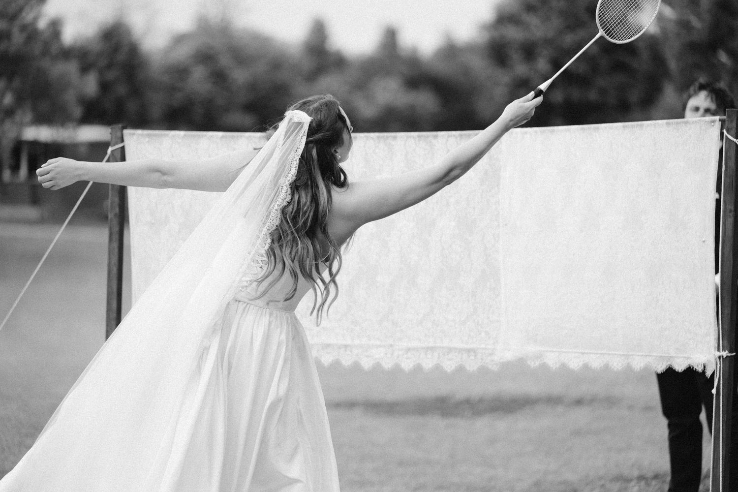 Bride in white dress and veil stretches arms wide near decorative lace backdrop outdoors.