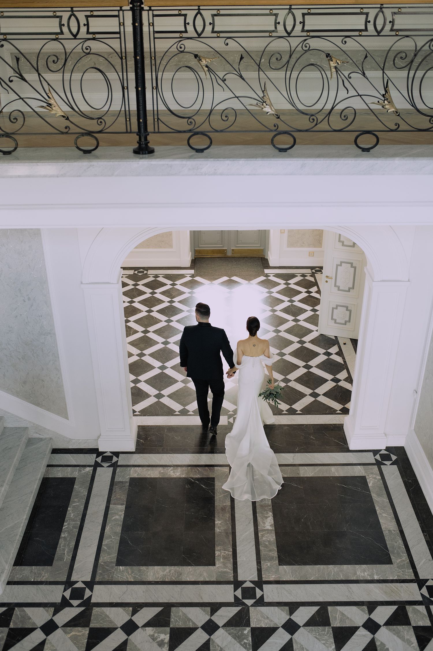 A couple in wedding attire walks across an elegant black and white marble floor with ornate railing details.