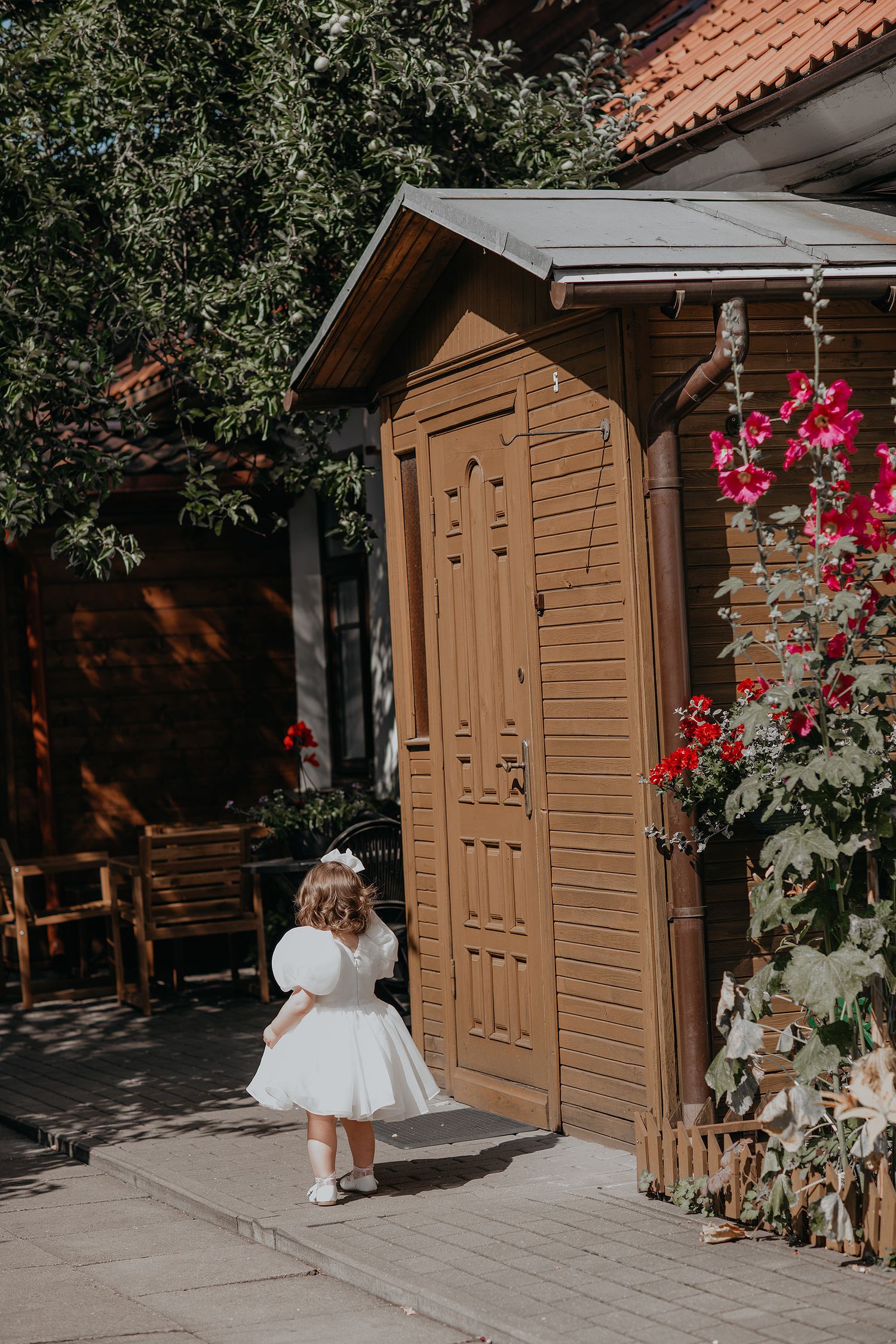 A small child in a white dress stands by a wooden door with pink flowers climbing on the wall of a rustic building.