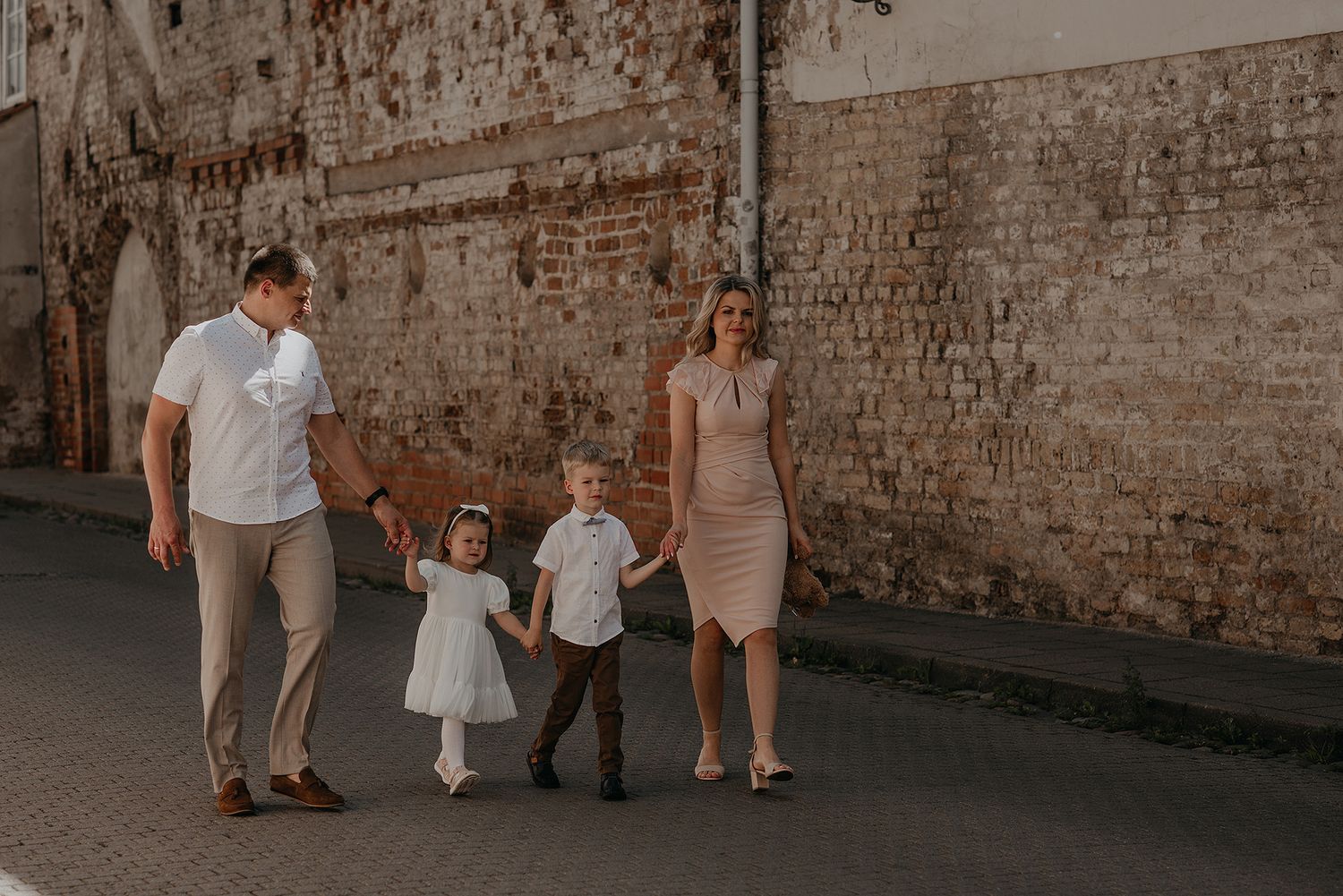 Family in coordinated neutral outfits walks along rustic brick wall during outdoor photo session.