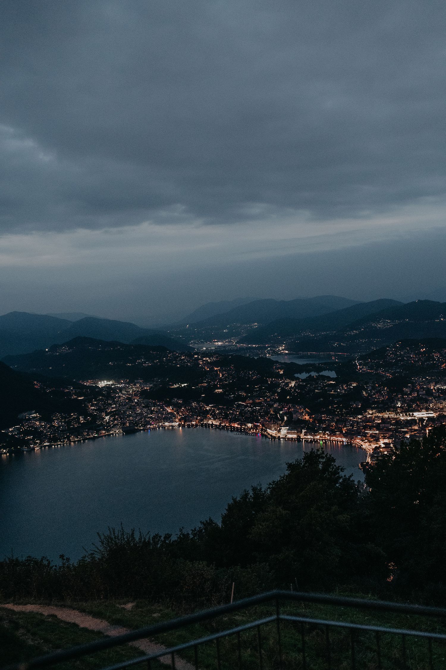 Panoramic dusk view of a coastal city with twinkling lights reflecting on the bay under moody cloudy skies.