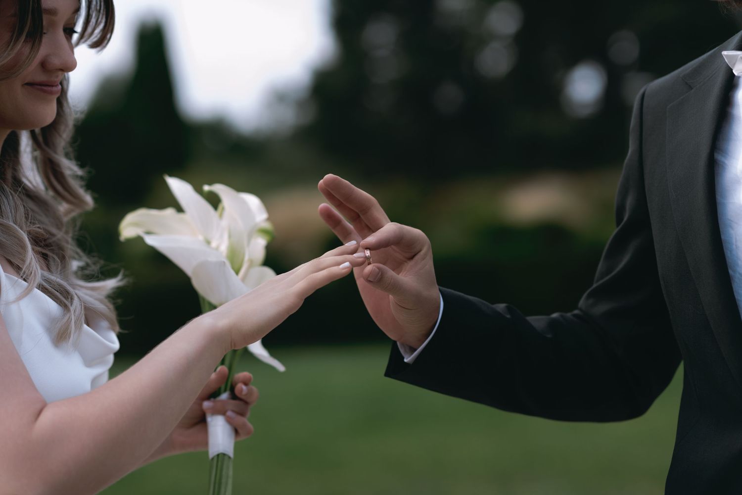 Bride in a white dress holding a white calla lily bouquet as the groom in a dark suit slides a wedding ring onto her finger outdoors.