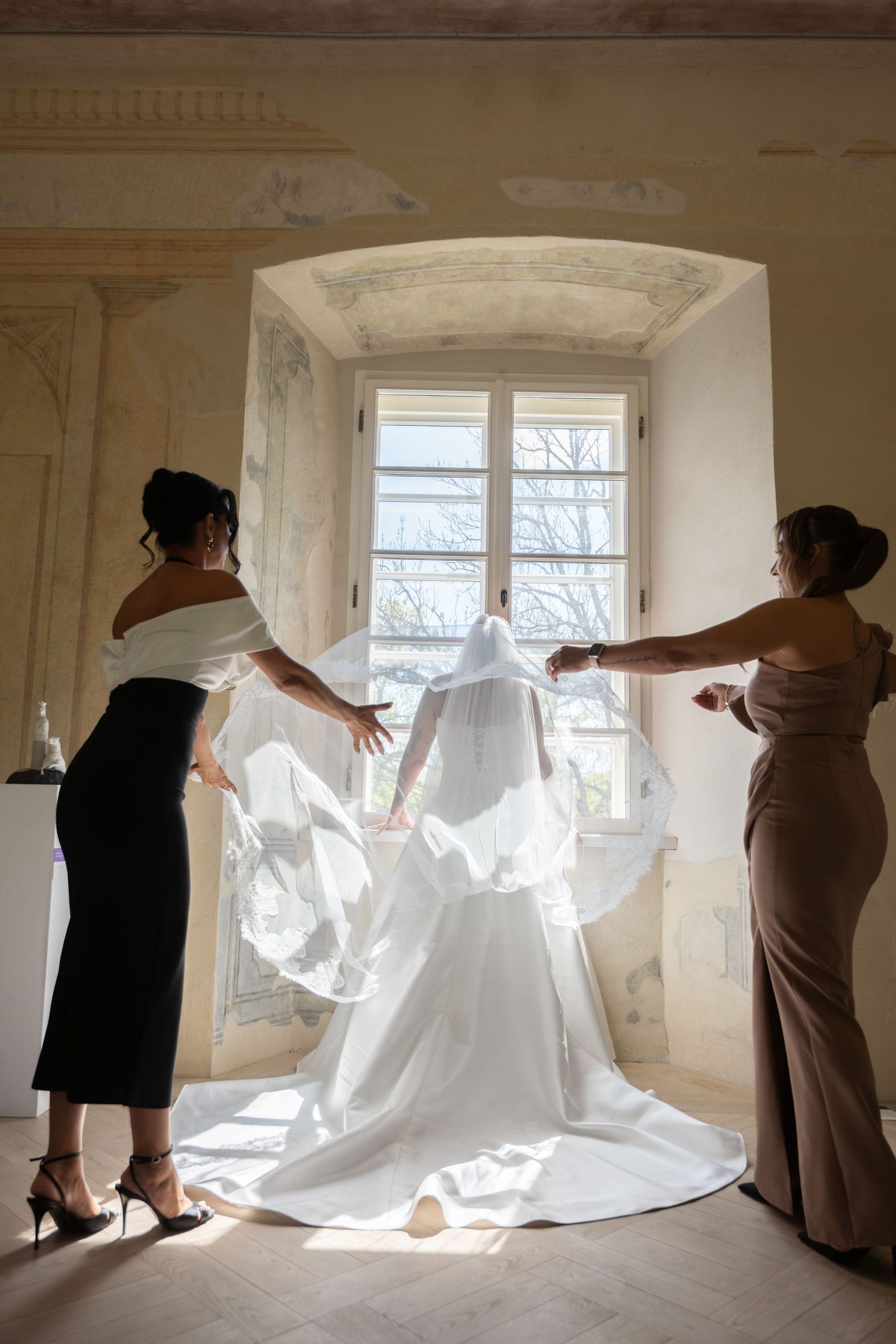 Two bridesmaids spread the bride's long veil and gown train as she faces a bright arched window in an elegant historic room.