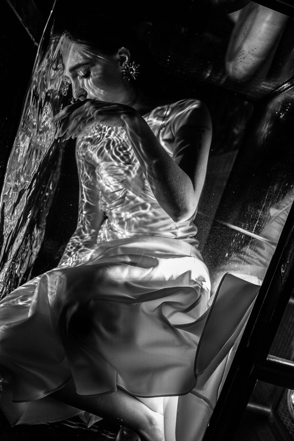 Black and white portrait of a woman in a satin dress lying on glass, with rippling water reflections patterning her back.