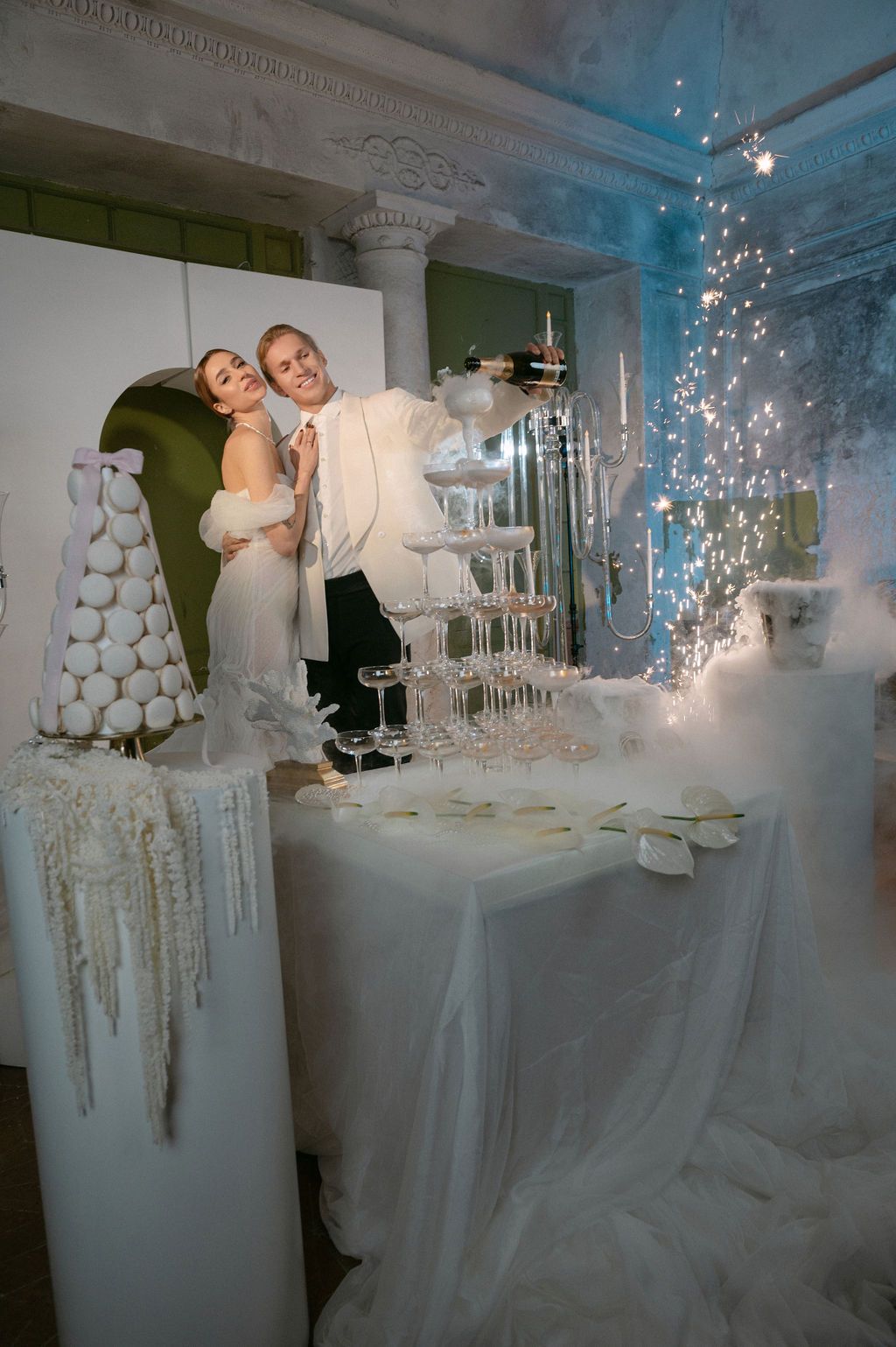 Bride and groom pose beside champagne tower and macaron display at elegant white wedding reception.