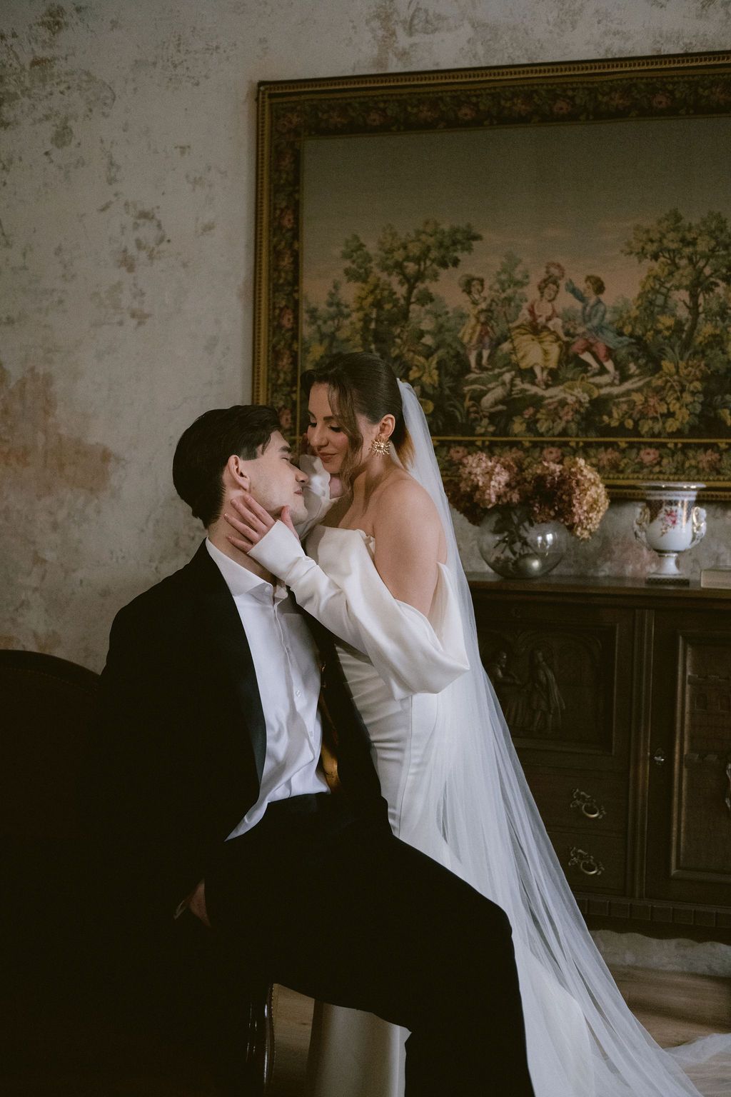 Smiling bride in off-shoulder white dress with veil tenderly holds grooms face in romantic vintage room.