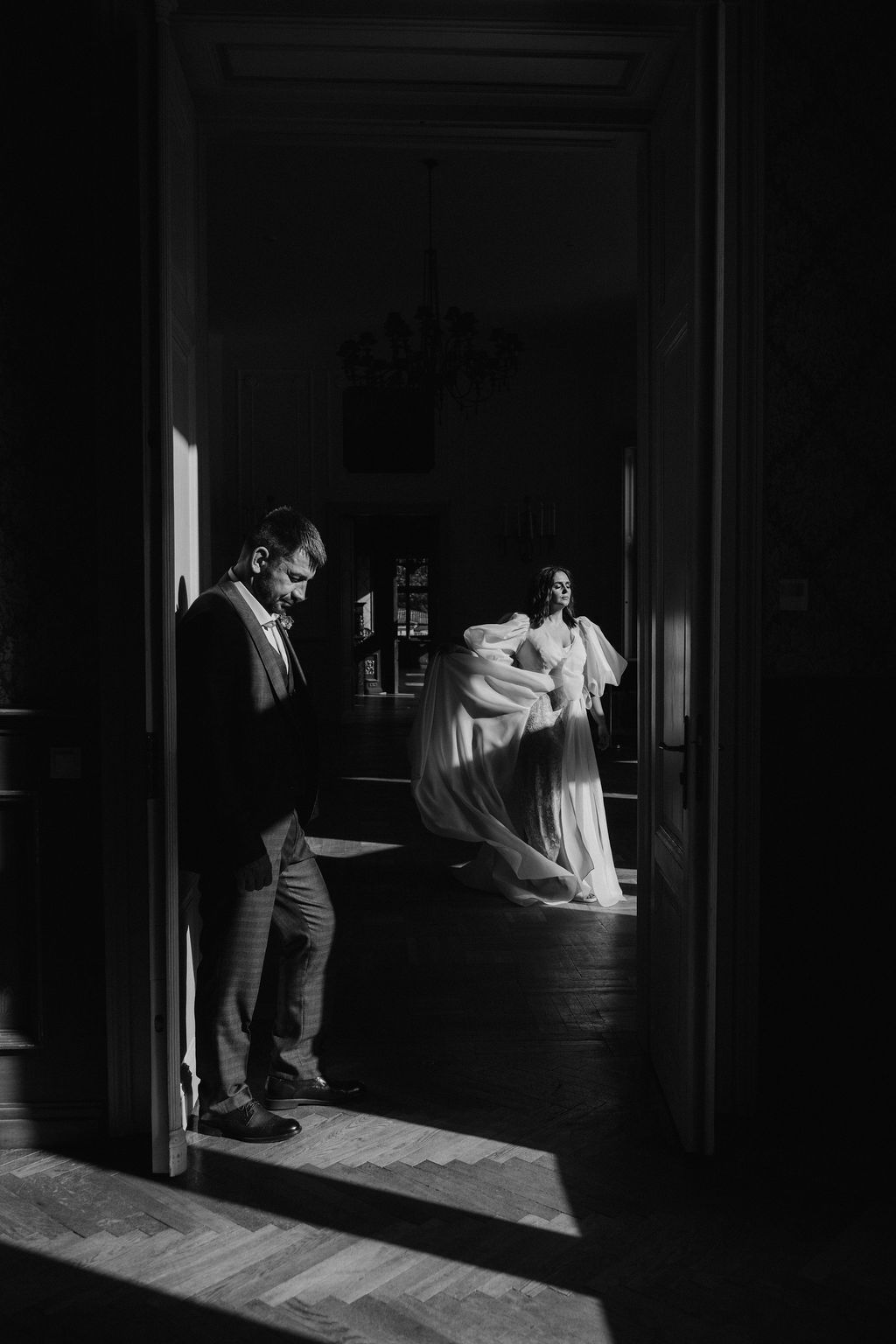 Black and white photo of groom leaning on doorframe while bride in flowing gown stands in sunlit room beyond.
