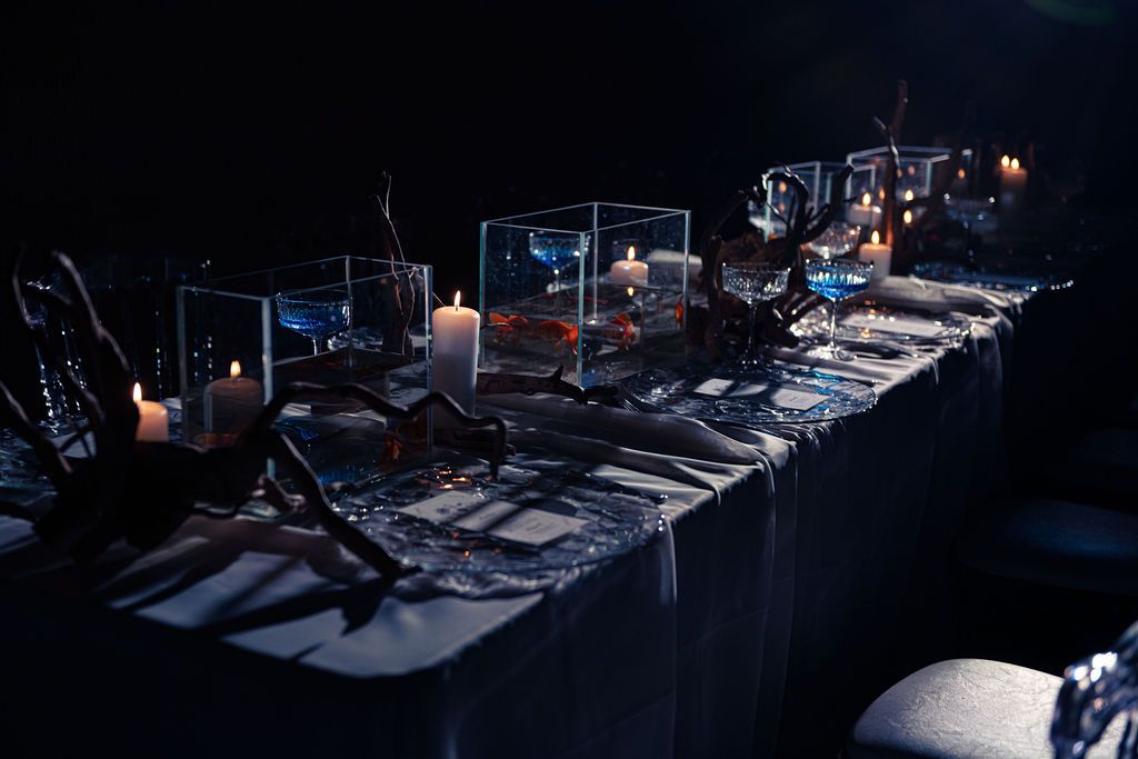 Elegant dark reception table with glowing candles, glass terrariums, and moody blue lighting for a sophisticated event.