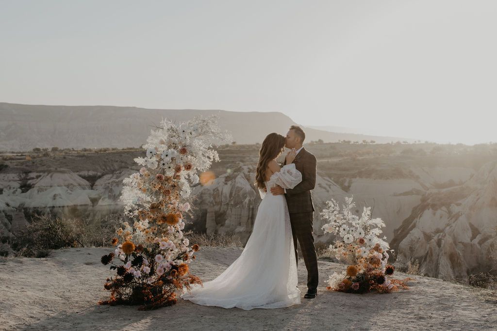 Couple kissing at outdoor wedding ceremony with boho floral arrangements in rocky desert landscape at golden hour.