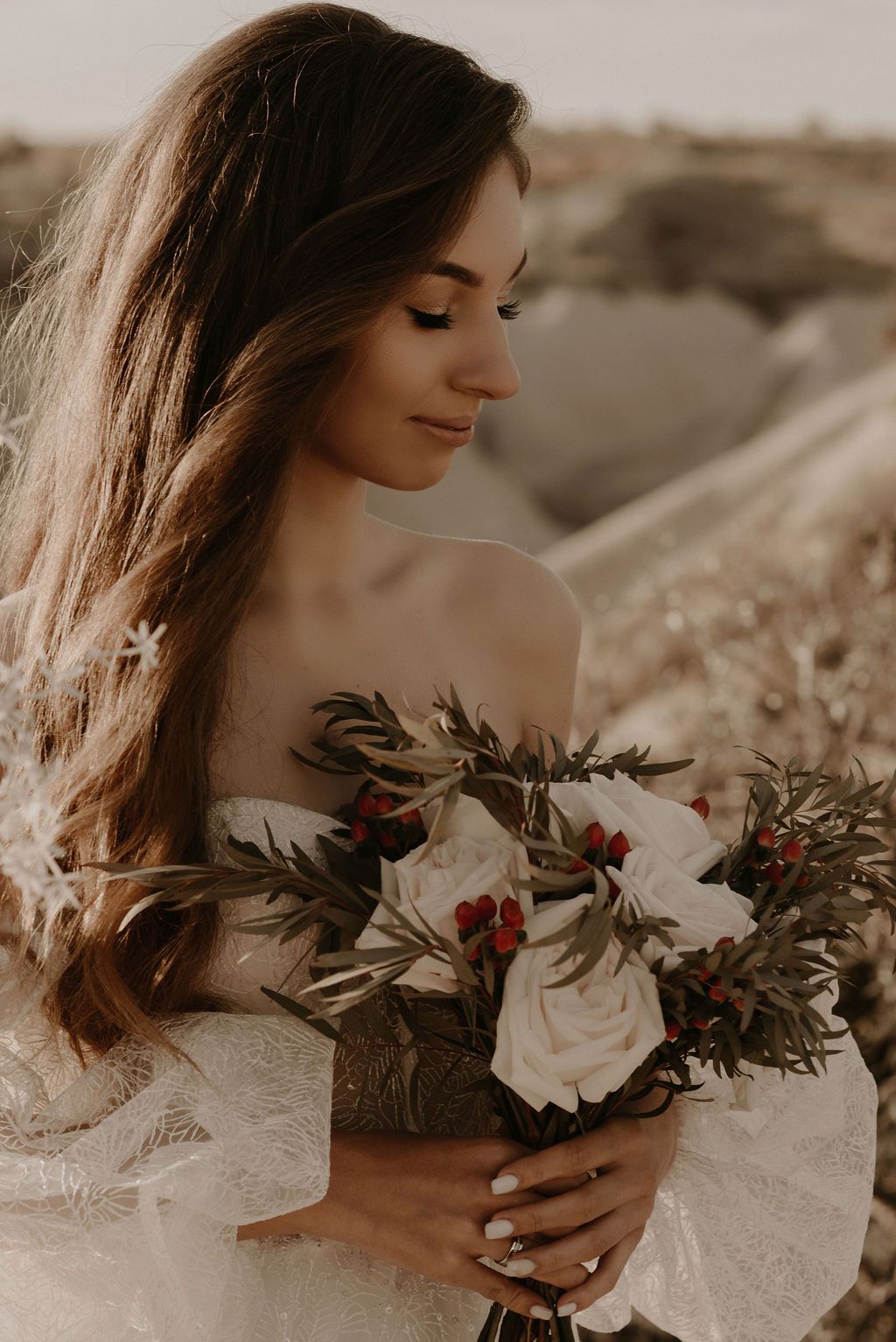 Bride in off-shoulder lace gown holding white rose and red berry bouquet with greenery outdoors.