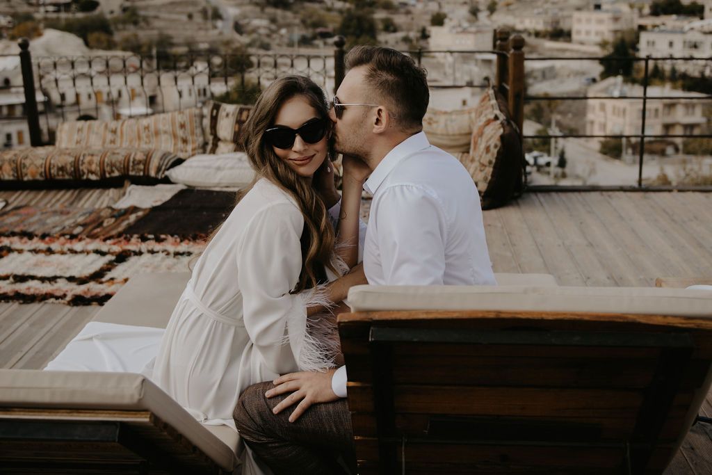 Couple sitting together on a rooftop terrace, woman in sunglasses and white outfit, man in white shirt kissing her cheek.