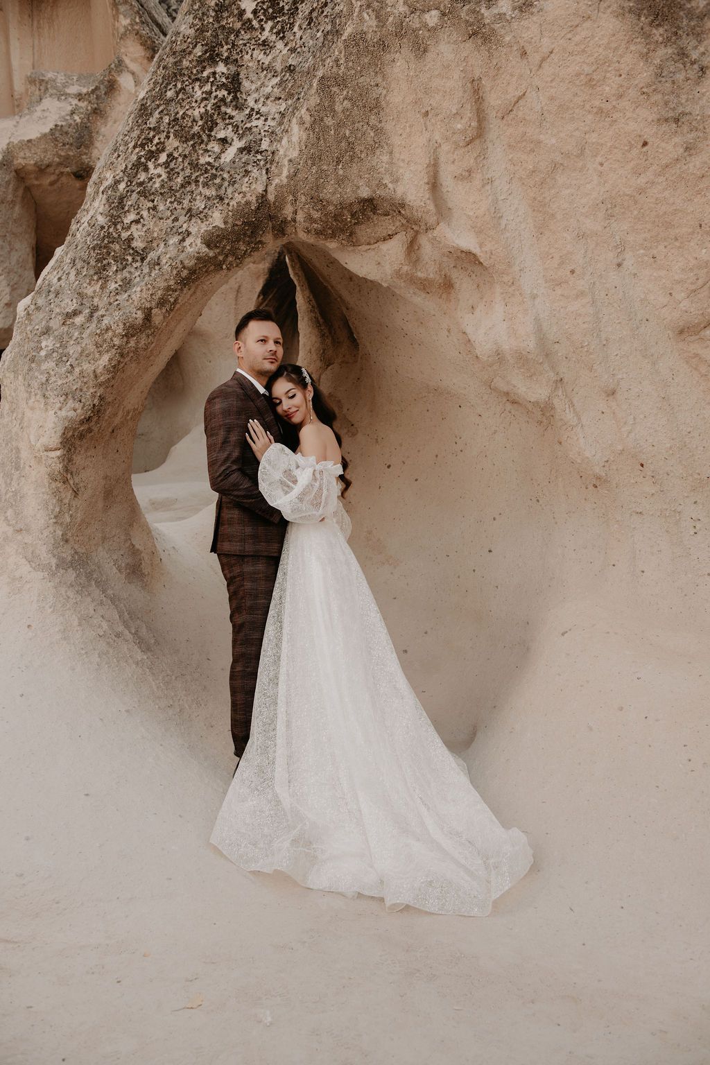 A bride and groom pose romantically in a white wedding dress and brown suit among ancient cave rock formations.