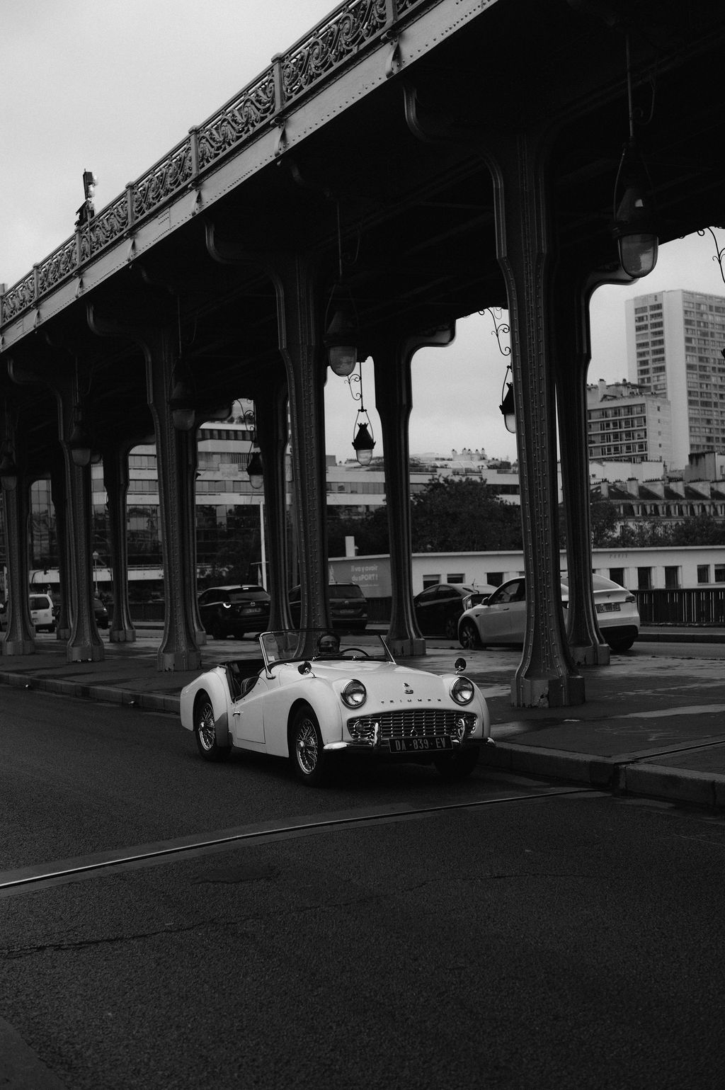 Vintage white convertible sports car parked under ornate Parisian bridge in black and white photo.
