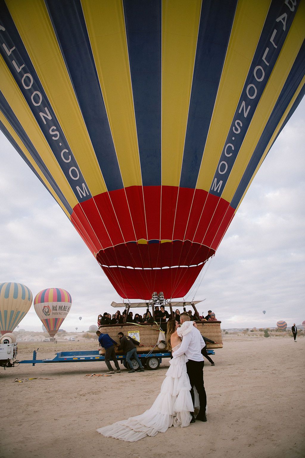 Bride and groom embrace beneath a colorful hot air balloon in Cappadocia, Turkey, with other balloons visible in the background.