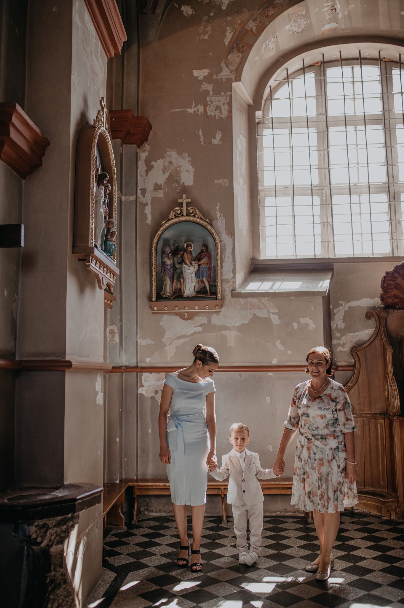 Woman and child walk with elderly lady inside a historic church with religious artwork and checkered floors.