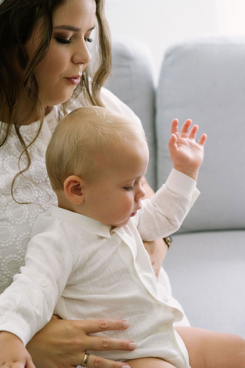 A mother tenderly holds her baby on her lap, the infant raising one hand while sitting in a bright living room.