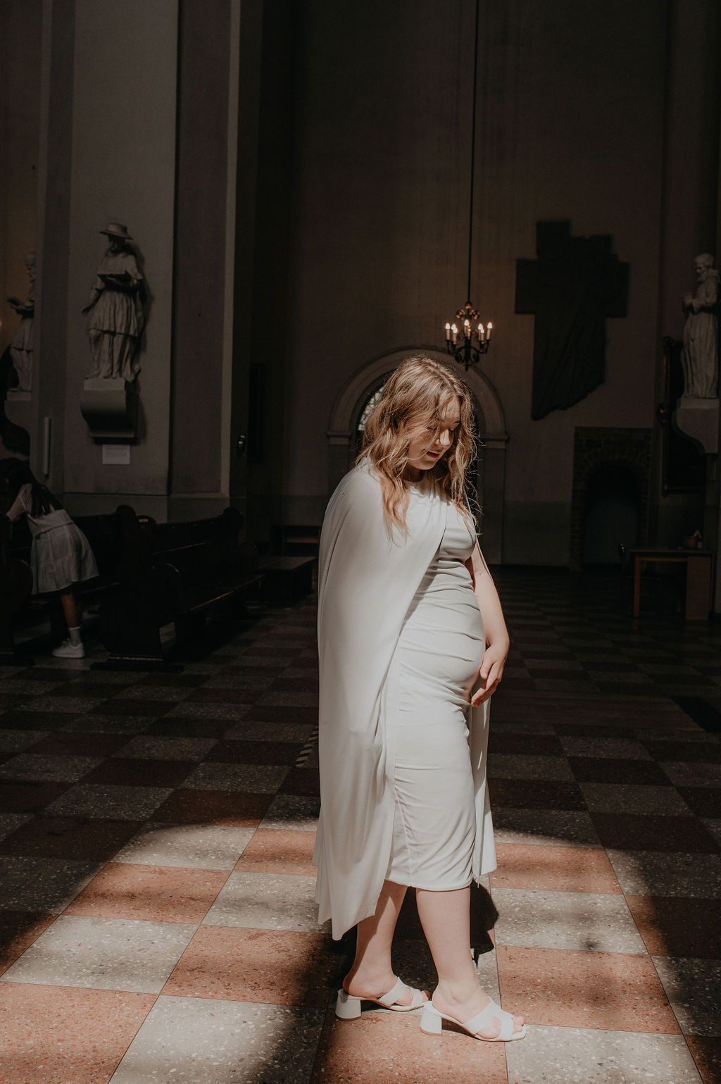 Pregnant woman in white dress and cape standing in church, looking down at baby bump in soft natural light.