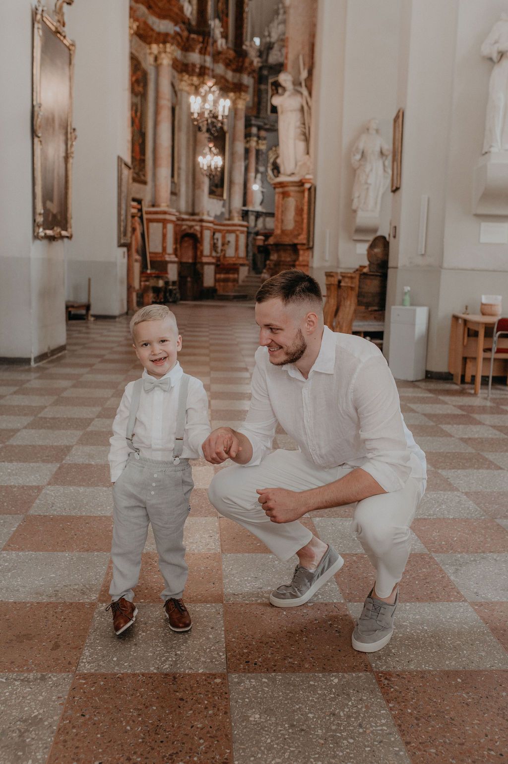 A father crouches beside his smiling toddler son in a elegant church, both dressed in white formal attire.