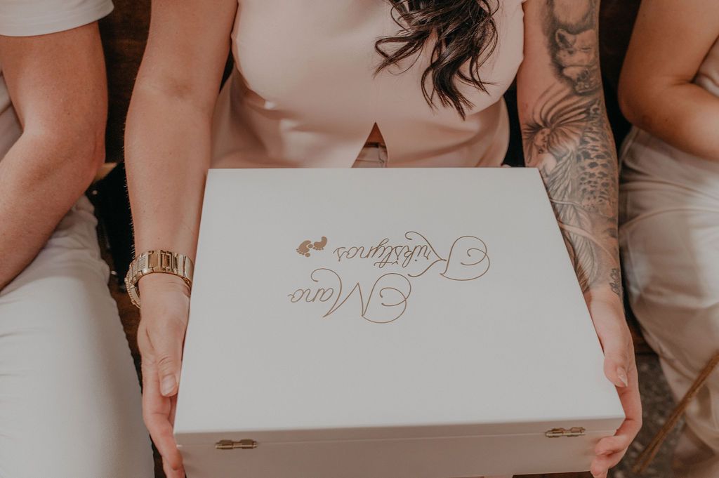 Woman holding a white personalized gift box with elegant script logo, wearing a pink top and gold bracelet.