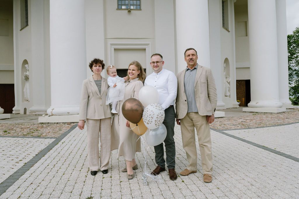 Family of five posing with balloons outside white columned building on paved courtyard.