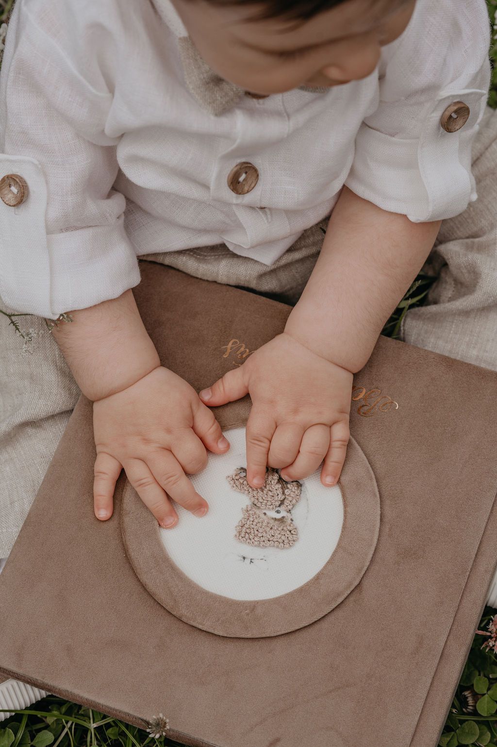 Baby in white linen shirt touching a illustrated bear print on a kraft paper book outdoors.