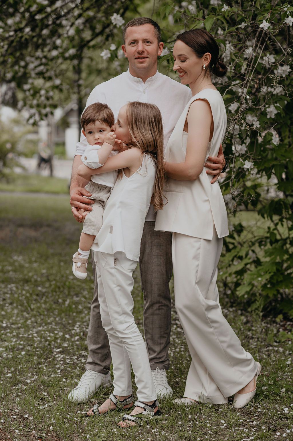 A happy family of four poses outdoors in white outfits near blooming trees on a sunny day.