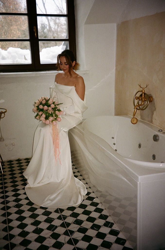 Bride in white strapless gown holding pink rose bouquet standing by jacuzzi tub on checkered tile floor.