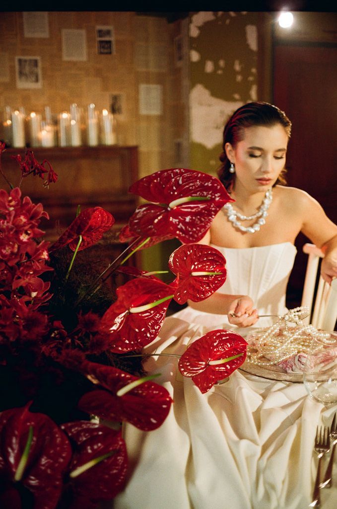 Bride in strapless white gown seated at elegant table with red anthurium flowers and pearl necklace in candlelit setting.