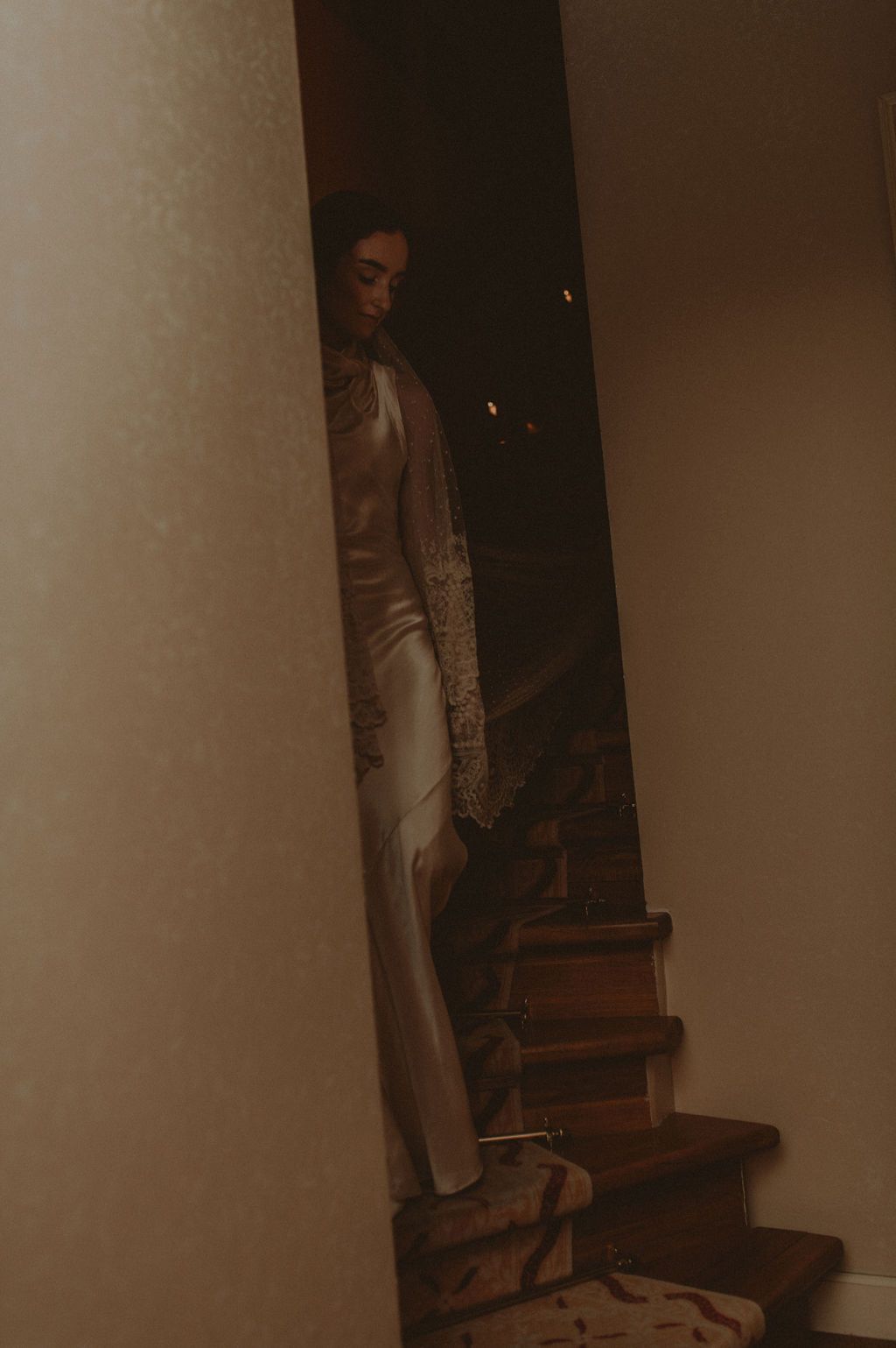 Bride in elegant white gown stands on wooden stairs in a dimly lit interior, captured through a narrow doorway.