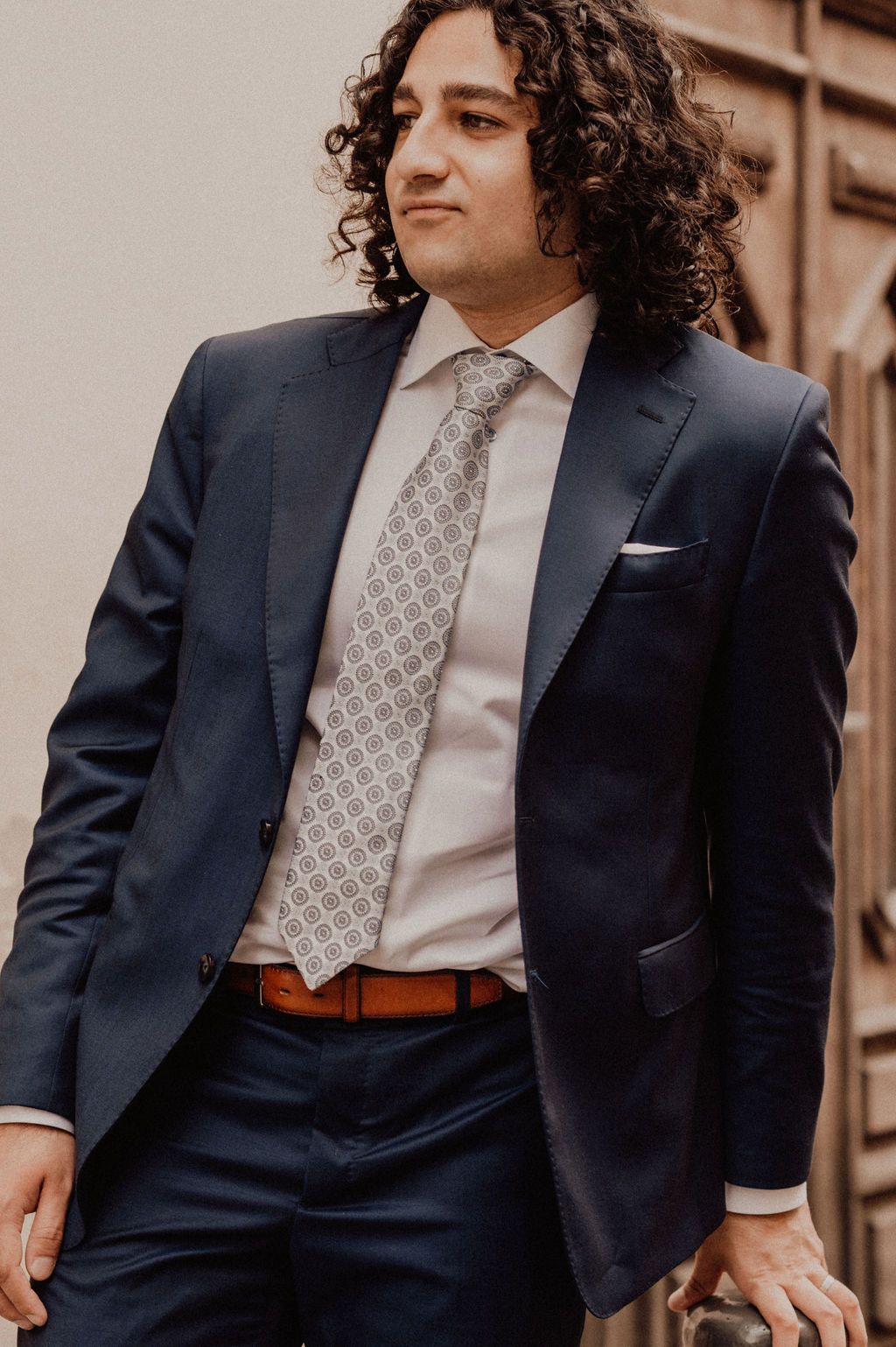 Man in navy suit with patterned tie and white pocket square, wearing brown belt, with curly hair.
