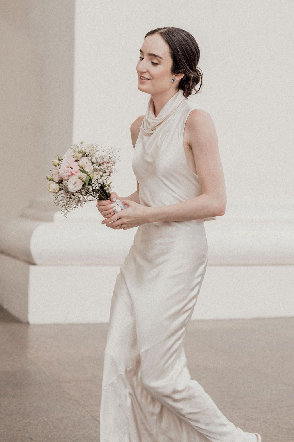 Elegant bride in a sleeveless ivory satin gown holding a pink and white bouquet, smiling softly outdoors.